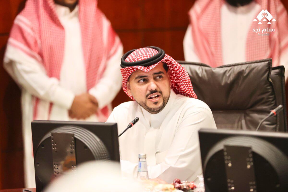 اجتماع رسمي لشخص يرتدي الثوب السعودي والشماغ، يتحدث أمام شاشة عرض مع وجود أشخاص آخرين في الخلفية.