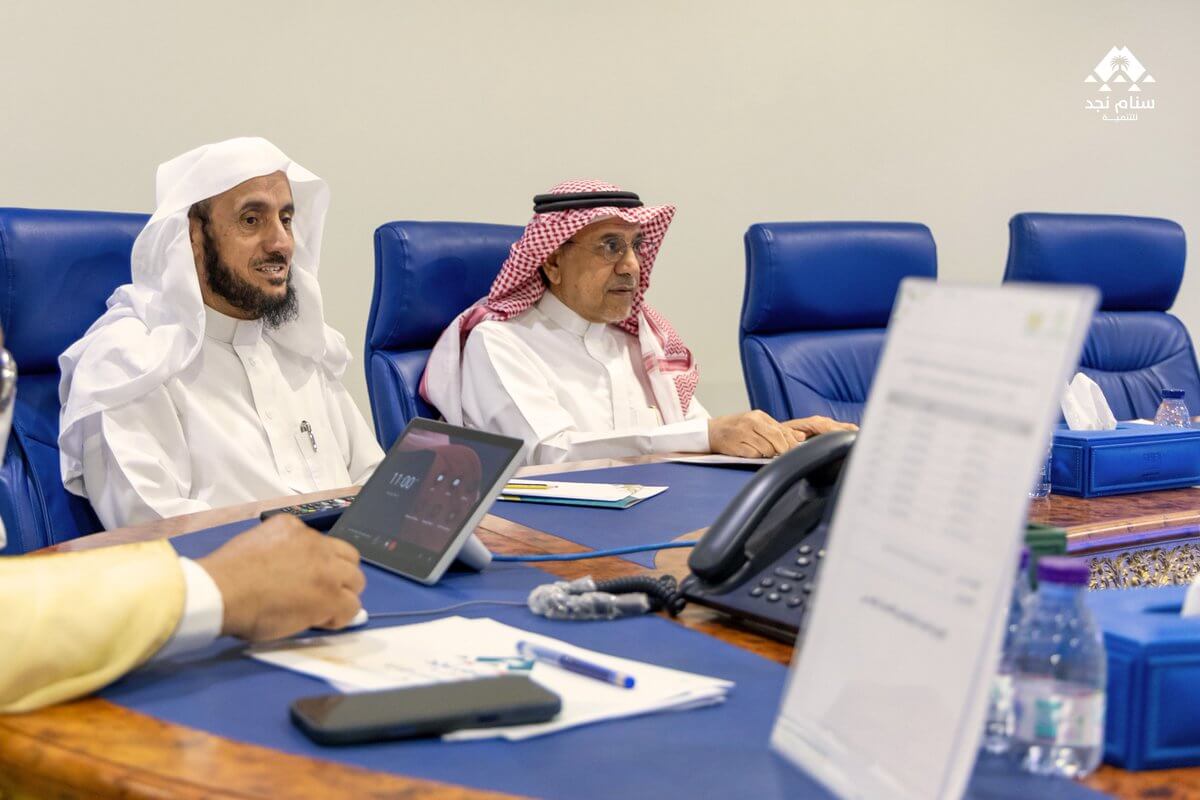 صورة لرجال يجلسون في اجتماع، يرتدون ملابس تقليدية عربية، مع وجود هواتف تعمل وأوراق على الطاولة.